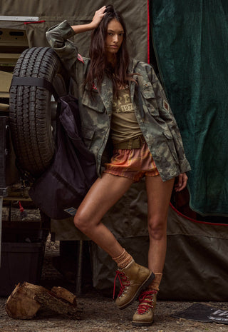 A woman poses in a rustic outdoor camping setting, wearing a camouflage utility jacket, a graphic t-shirt, orange shorts, and tan lace-up boots, with a large spare tire and tent visible in the background.