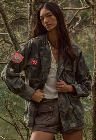 A woman wearing a military-style camouflage jacket with red patches, a white top, and brown shorts, standing in a wooded outdoor setting.