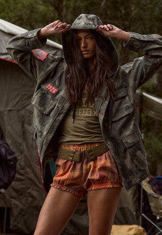 A woman wearing a vintage-style military camouflage jacket over an olive green graphic t-shirt, paired with orange plaid shorts and a wide belt, posing in front of a tent outdoors.