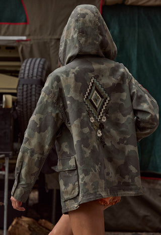 A person viewed from behind wearing a hooded camouflage jacket with a large geometric diamond pattern and small white flower details on the back, set against a dark outdoor backdrop.