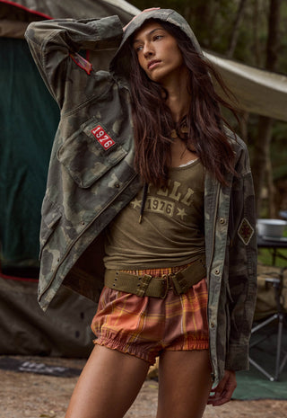 A woman wearing a vintage-style olive green camouflage jacket with a red '1976' patch, an olive green graphic tank top, and orange-brown plaid shorts, posed in front of a rustic outdoor tent.