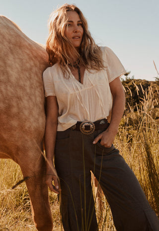 A woman stands outdoors in a field next to a horse, wearing a cream short-sleeve blouse tucked into dark brown wide-leg trousers with a western-style belt and large buckle.