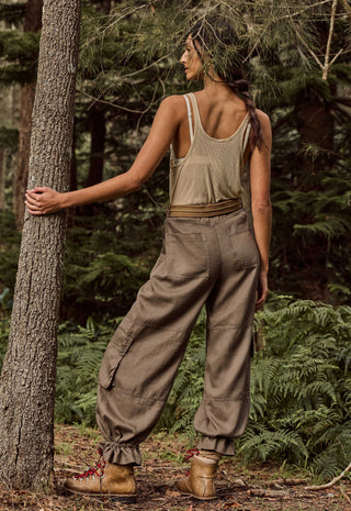 A woman stands in a forest wearing a sleeveless beige tank top, loose brown cargo pants, and brown hiking boots, with her back turned and one hand resting on a tree trunk.