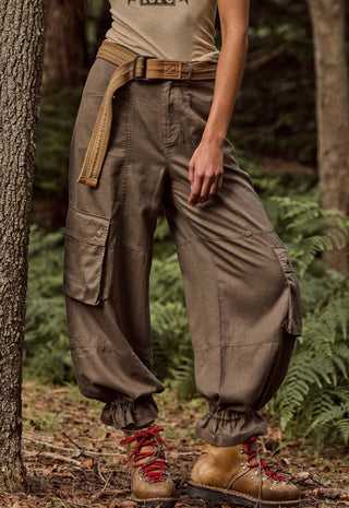 A person standing outdoors in a forest wearing loose brown cargo pants with gathered cuffs, a tan belt, and brown lace-up boots.
