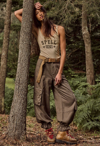 A woman posing in a forest setting, wearing a beige tank top with 'SPELL 1976' printed on the front, baggy brown cargo pants, a tan belt, and brown hiking boots.