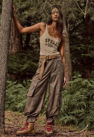 A woman stands in a forest wearing a beige tank top with 'SPELLS 1976' printed on it, loose brown cargo pants with a matching belt, and brown hiking boots with red laces.