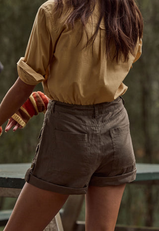 A person shown from the back wearing olive brown utility shorts with rolled cuffs and a tan button-up shirt, standing outdoors against a blurred forest background.