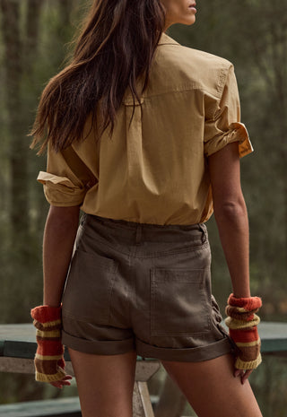 A woman shown from the back wearing a tan short-sleeve button-up shirt and brown shorts, with striped knit fingerless gloves, standing outdoors against a blurred forest background.