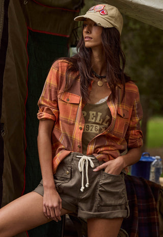 A woman wearing a tan baseball cap, an orange and brown plaid flannel shirt, a brown graphic tank top, and olive cargo shorts, posed in front of a tent in an outdoor setting.