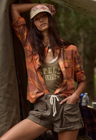 A woman posing in a rustic outdoor setting, wearing a rust-orange and yellow plaid flannel shirt over a graphic tank top, olive green cargo shorts, and a beige baseball cap.