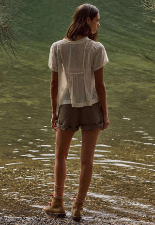 A woman stands at the edge of shallow water, viewed from behind, wearing a loose white short-sleeve blouse, olive-brown shorts, and tan ankle boots.