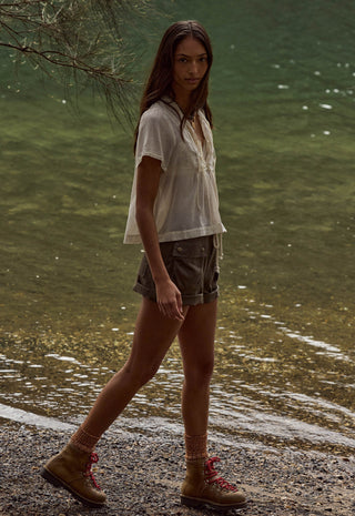 A woman walking along a rocky shoreline next to a body of water, wearing a loose white short-sleeve top, dark gray shorts, and tan hiking boots with red laces.