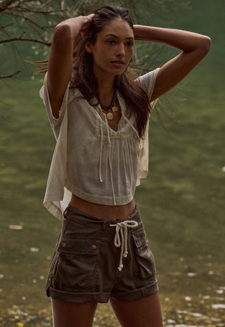 A woman standing outdoors near water, wearing a loose short-sleeve cream top and dark brown cargo shorts with a white drawstring.