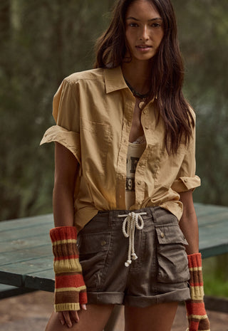 A woman wearing a tan button-up shirt, brown cargo shorts, and striped knit fingerless gloves, standing outdoors in front of a blurred forest background.