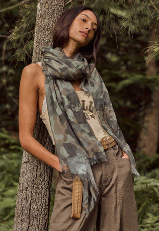 A woman in a forest setting wearing a camouflage print scarf draped over her shoulders, paired with a beige tank top and brown cargo pants.
